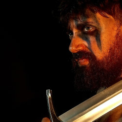A white man holding a Viking sword, with black face paint across his left eye and cheek