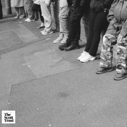 Grayscale image of 6 people's legs. One person has an assisted wheeled frame. Three are wearing black trousers, two are wearing grey trousers and one is wearing camouflaged trousers.