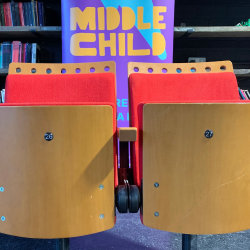 Two wooden theatre seats with red fabric in front of a Middle Child banner.