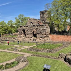 A photo of the Jewry Wall Museum on a sunny day. St Nicholas Church is behind the wall and the rest of the Baths ruins is in front. There are people walking around the ruins.