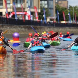 A group of kayakers paddle brightly coloured boats along a calm river, wearing red helmets and life jackets.