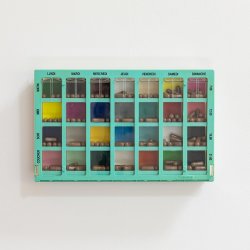 A bright turquoise weekly pill organiser mounted on a white wall holds brown capsules. Each compartment for the days of the week features a different coloured background. The writing is in French.