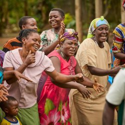Women coffee farmers from Bukonzo, Uganda are dancing and singing. They are smiling and have colourful clothes on. Beige, Green, Pink and Yellow, Blue and Red stripes,