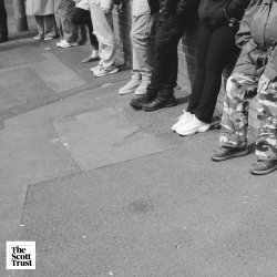 Grayscale image of 6 people's legs. One person has an assisted wheeled frame. Three are wearing black trousers, two are wearing grey trousers and one is wearing camouflaged trousers.