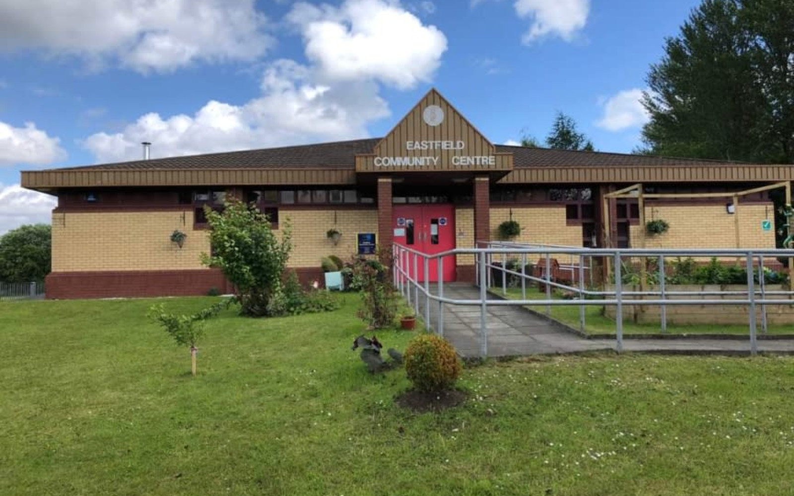 Exterior of Eastfield Community Centre, a single-story tan and red brick building with a peaked roof sign above red double doors, entrance ramp, and surrounding greenery under a partly cloudy sky.