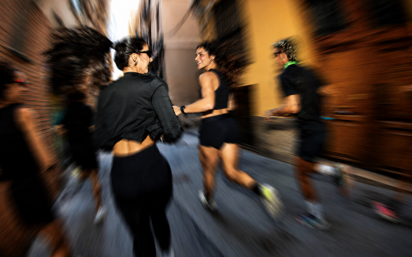 A group of runners in black athletic clothing moving quickly through a narrow city alley, captured with strong motion blur to convey speed and energy.