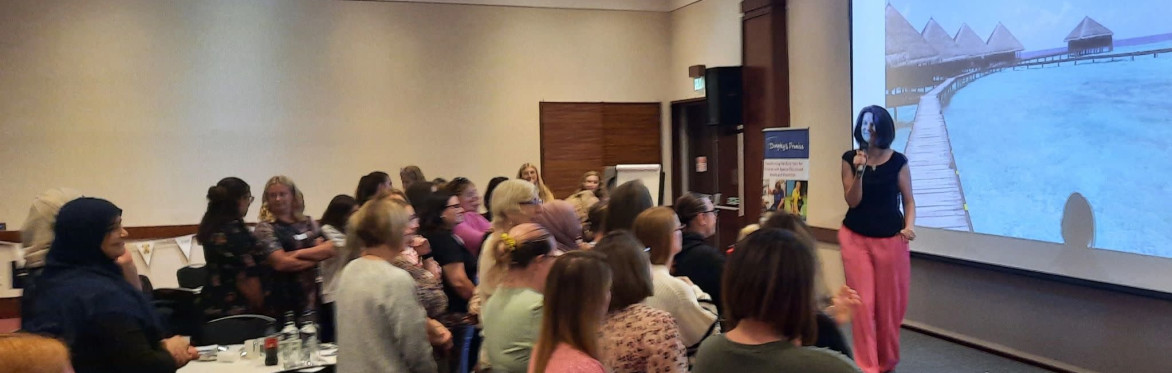 Event with lots of people and a woman with brown hair, a black top and pink trousers speaking into a microphone