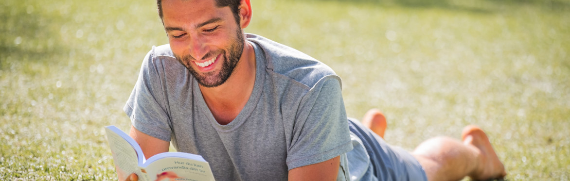 A smiling younger man with summer clothes lying on grass reading a book