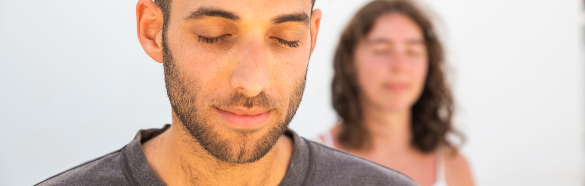 Two peaceful meditators, in the foreground a young man with short hair and a brown tee shirt, in the background a young woman with shoulder length hair and white top.