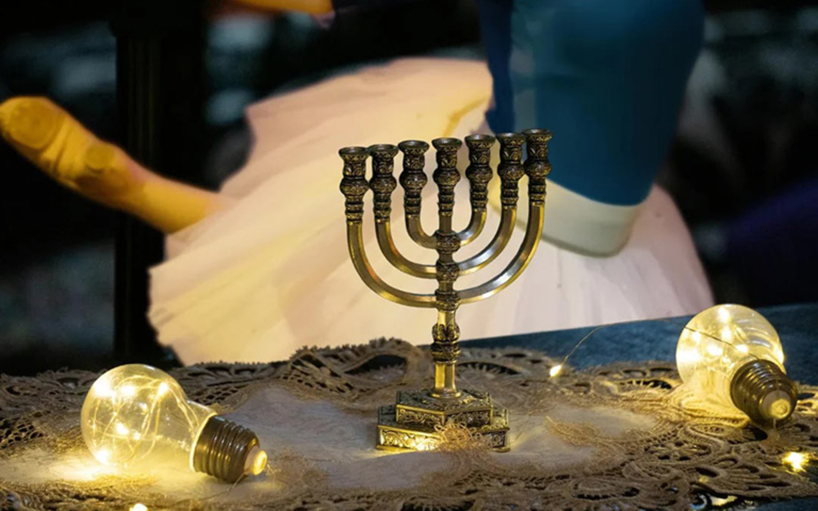 A picture of an unlit menorah on a table with an ornate tablecloth adorning a table is in focus, while an ambiently lit ballerina's tutu is in the background, worn by a mysterious dancer.