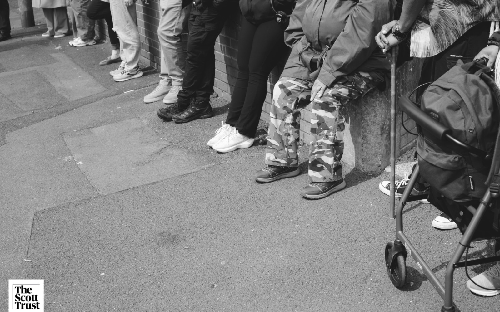 Grayscale image of 6 people's legs. One person has an assisted wheeled frame. Three are wearing black trousers, two are wearing grey trousers and one is wearing camouflaged trousers.