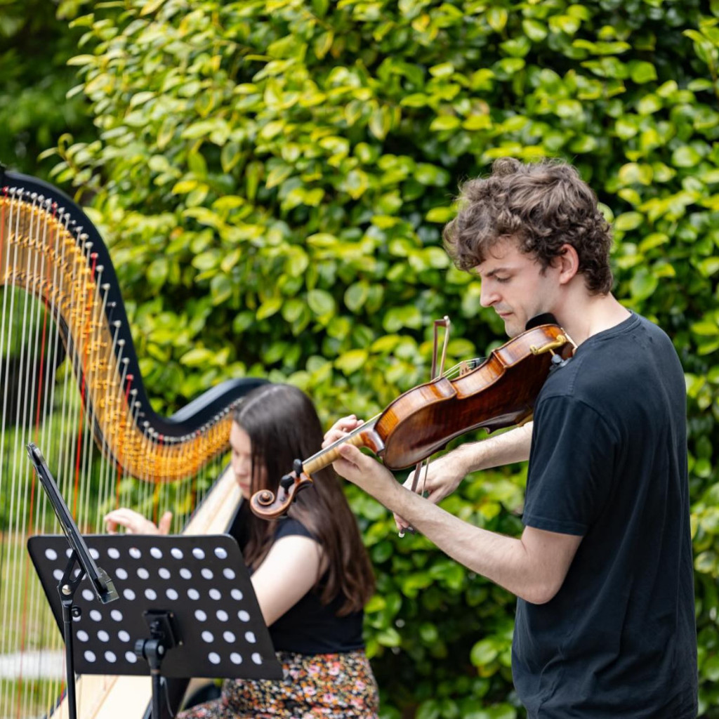James Warburton (violin, front) and Angharad Huw (harp, back) performing outside with a big leafy bush as a backdrop. They are both wearing black t-shirts and looking at their music.