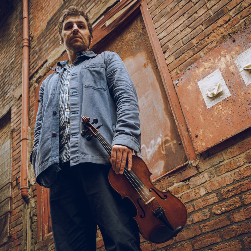 Low shot image of Mikey Kenney standing in front of an outside wall holding a fiddle.
