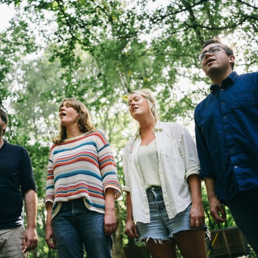 Four adults standing outdoors, looking up and singing among green trees.