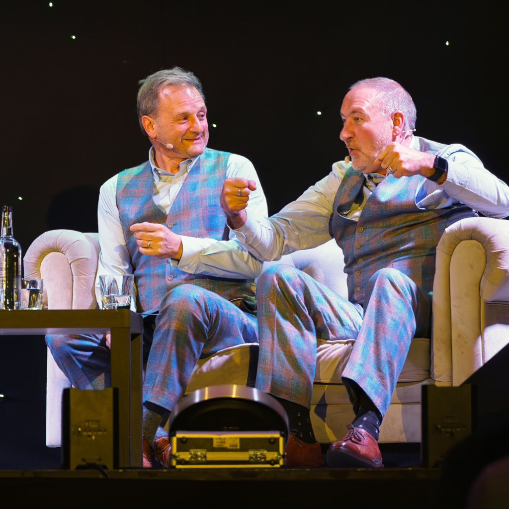 The image features Mark Radcliffe and Marc “Lard” Riley sat on a sofa on a stage in waistcoats, shirts and trousers, gesticulating with clenched fists
