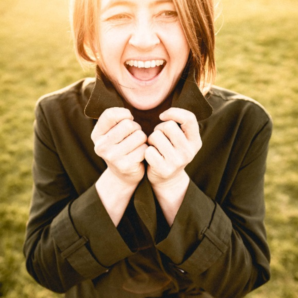 Karine Polwart laughing in a sunny field. Karine is wearing a dark green trench coat and holding on to the collar.