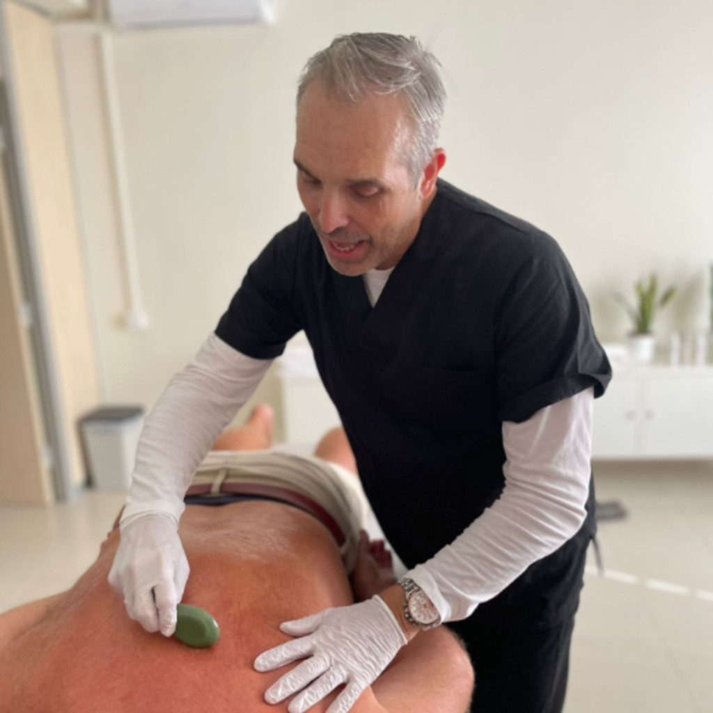 Clive Witham, a white male with grey short hair, giving a gua sha treatment to a patient on their back