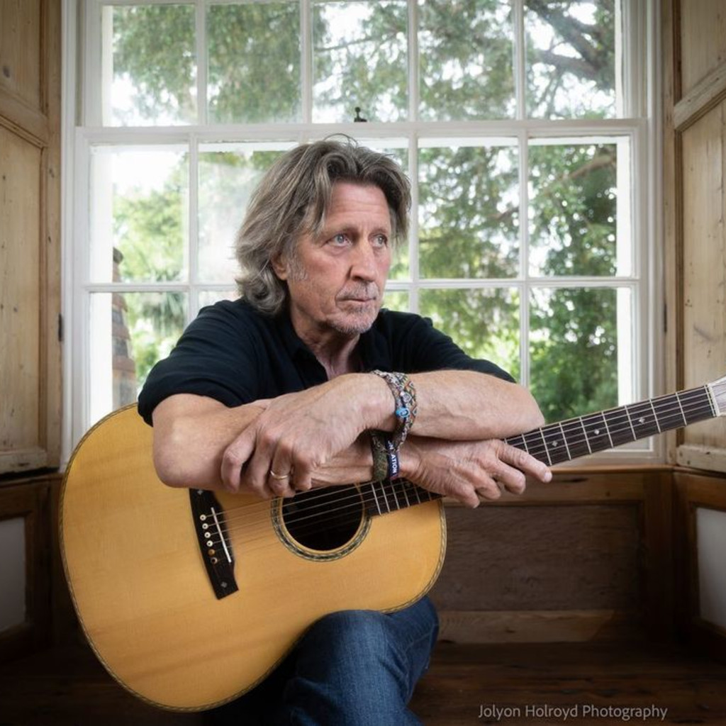 Steve Knightley, with mid length grey hair, sits in front of a window, posing with both arms folded over his guitar, looking off to the right.