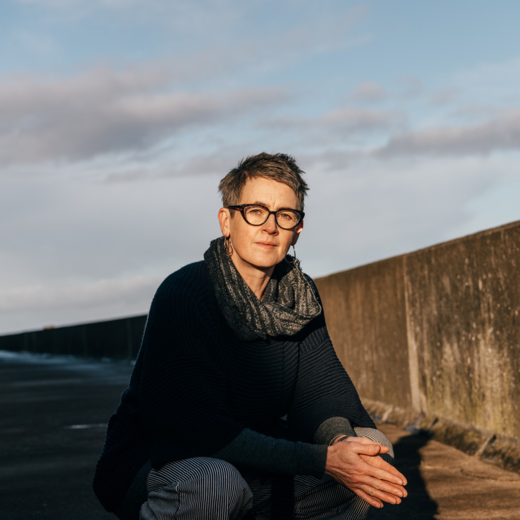 The image features Karine Polwart in a scarf and jumper squatting down in front of a low concrete wall with a dusky sky with whispy clouds behind her