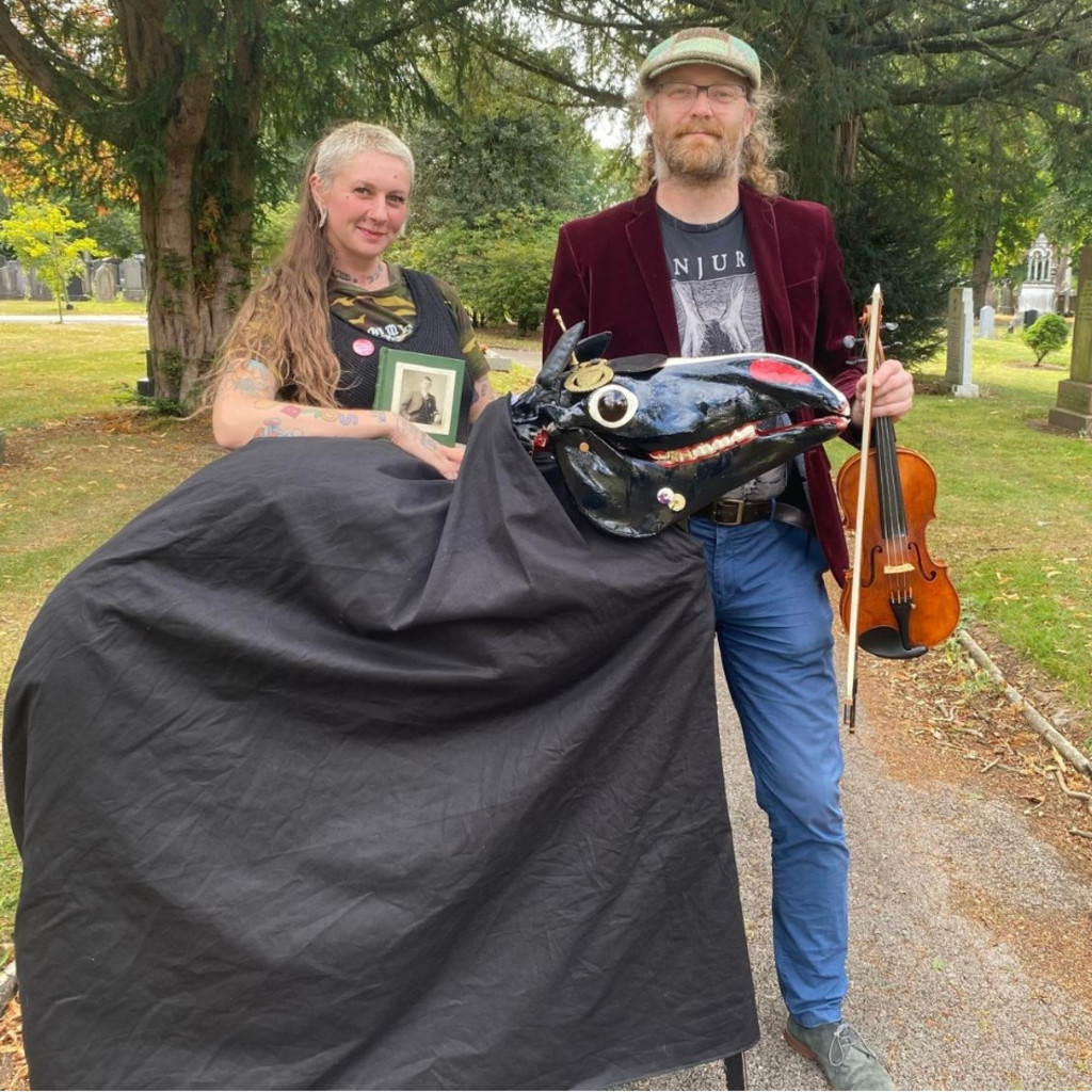 The image feature Jennifer Reid and Tom Kitching standing in a tree lined path behind a black, covered, skeletal horse. Tom is holding a fiddle. in his left hand.