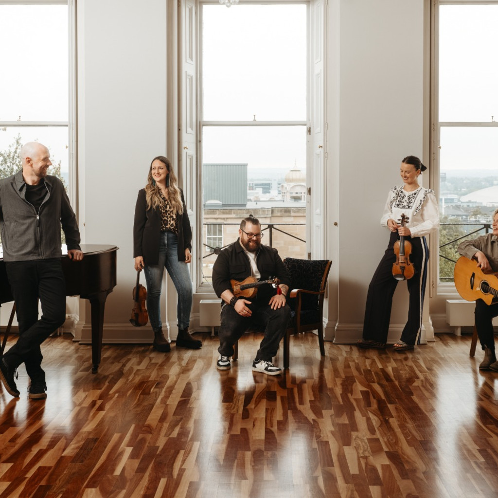 Five people of mixed gender, holding or standing with instruments, in a well lit, room with large windows.