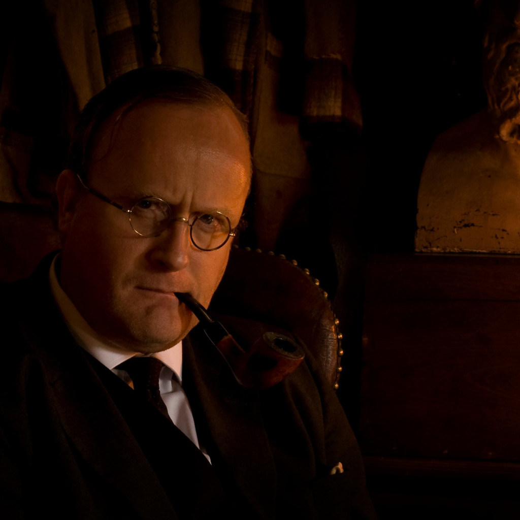 A portrait of Robert Lloyd Parry wearing a suit with a pipe in his mouth, sitting on a high backed leather chair