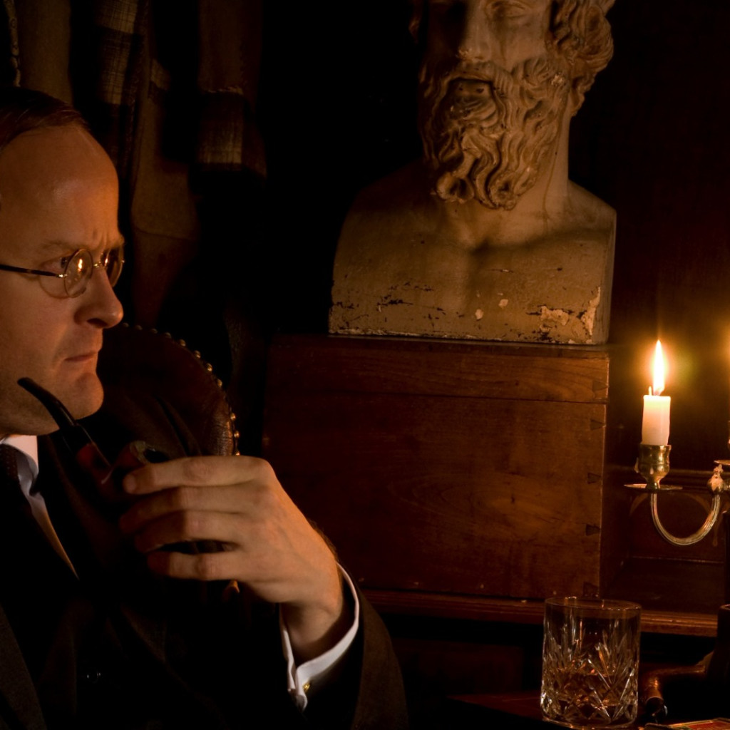 A side profile of Robert Lloyd Parry wearing a suit and holding a pipe in their left hand. To the right in the background are lit candlesticks and a bust of a man with a beard.