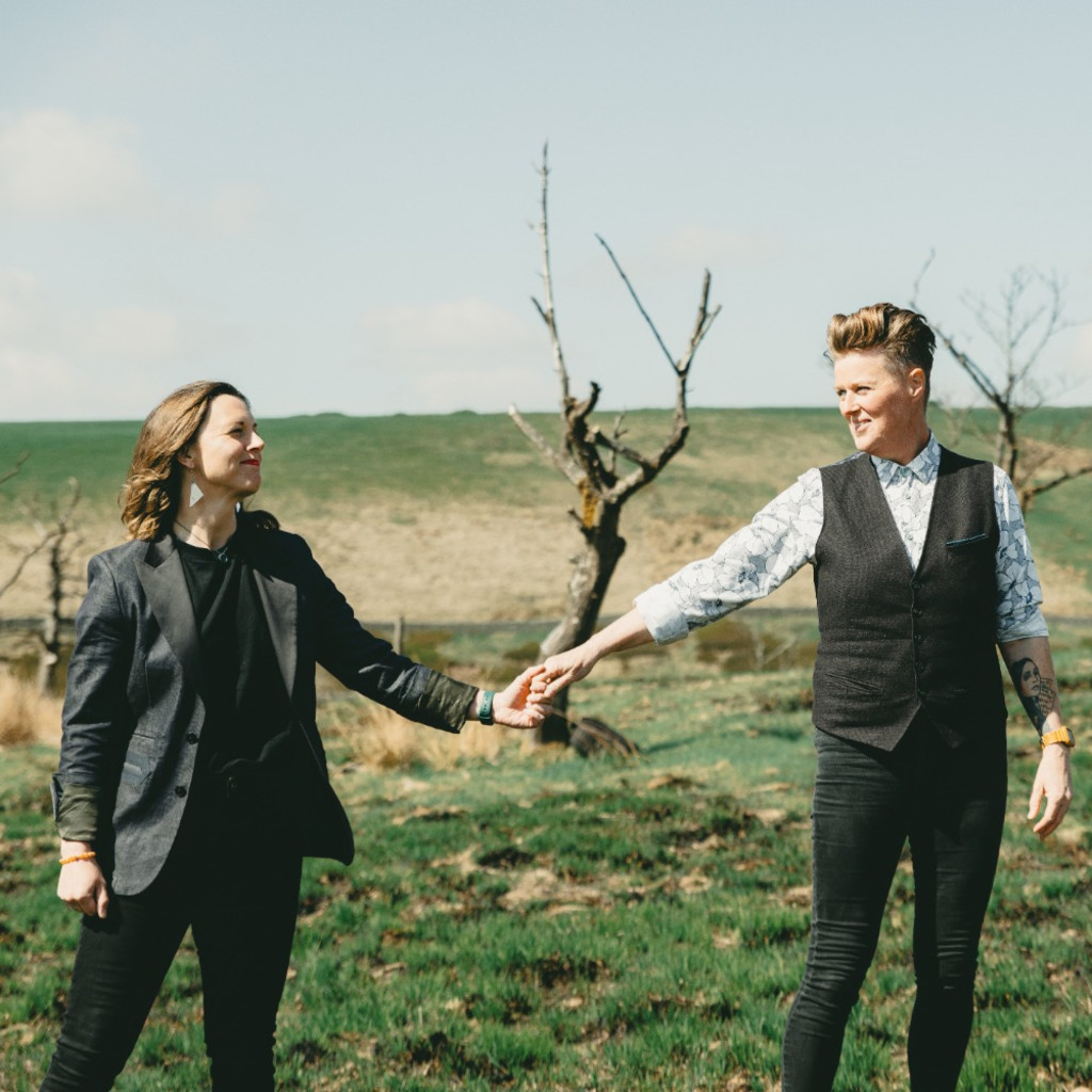 The image features two people holding hands, one has short blonde hair, and one has long brown hair. They are outside in an arid land with a bare tree behind them.