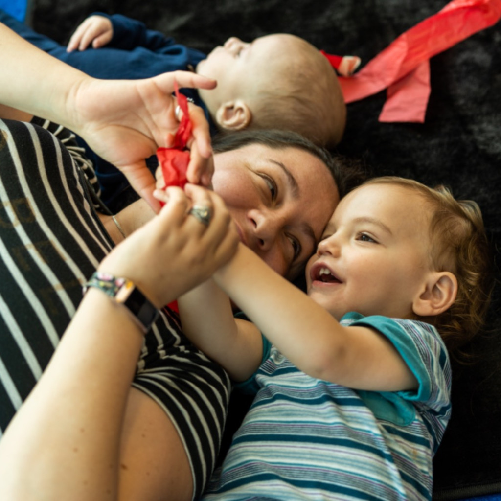 A young child and their carer  lying on a black mat and smiling. They are holding a piece of red paper tissue