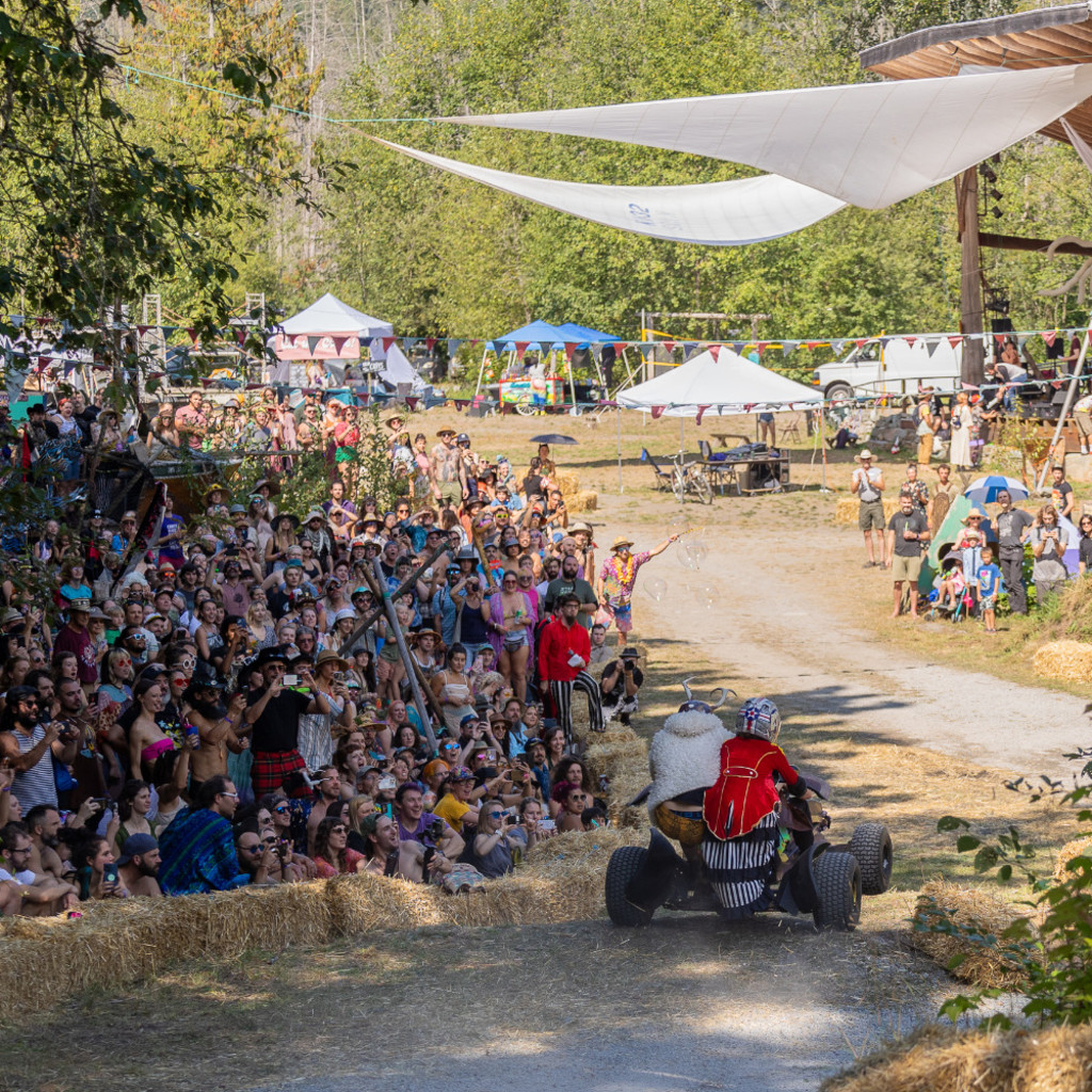 An outdoor scene of a crowd of cheering people on the left and a soapbox derby car with two costumed riders on the right
