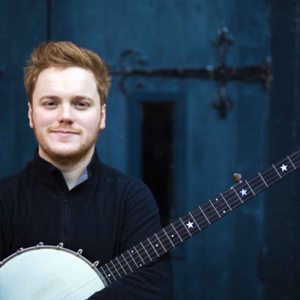 The image features Greg Russel facing the camera and smiling while holding a banjo. He is in front of a blue door with large gothic hinges.