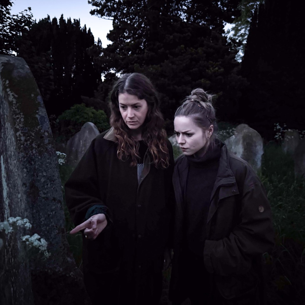 two girls standing infront of a gravestone in a dark churchyard and reading the text on the stone.