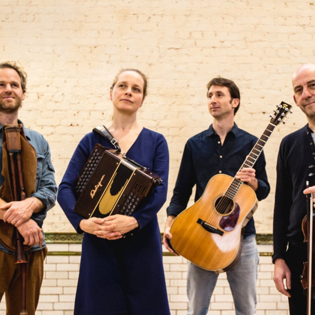 The image features Naragonia Quartet standing in front of a cream painted brick wall holding their instruments