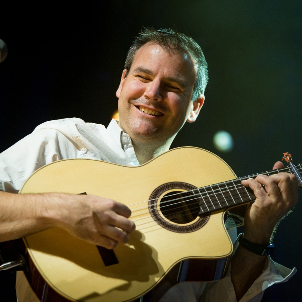 A close up of Tim Edey playing guitar on a dark stage. He is wearing a white shirt, his guitar is light wood and the background is black