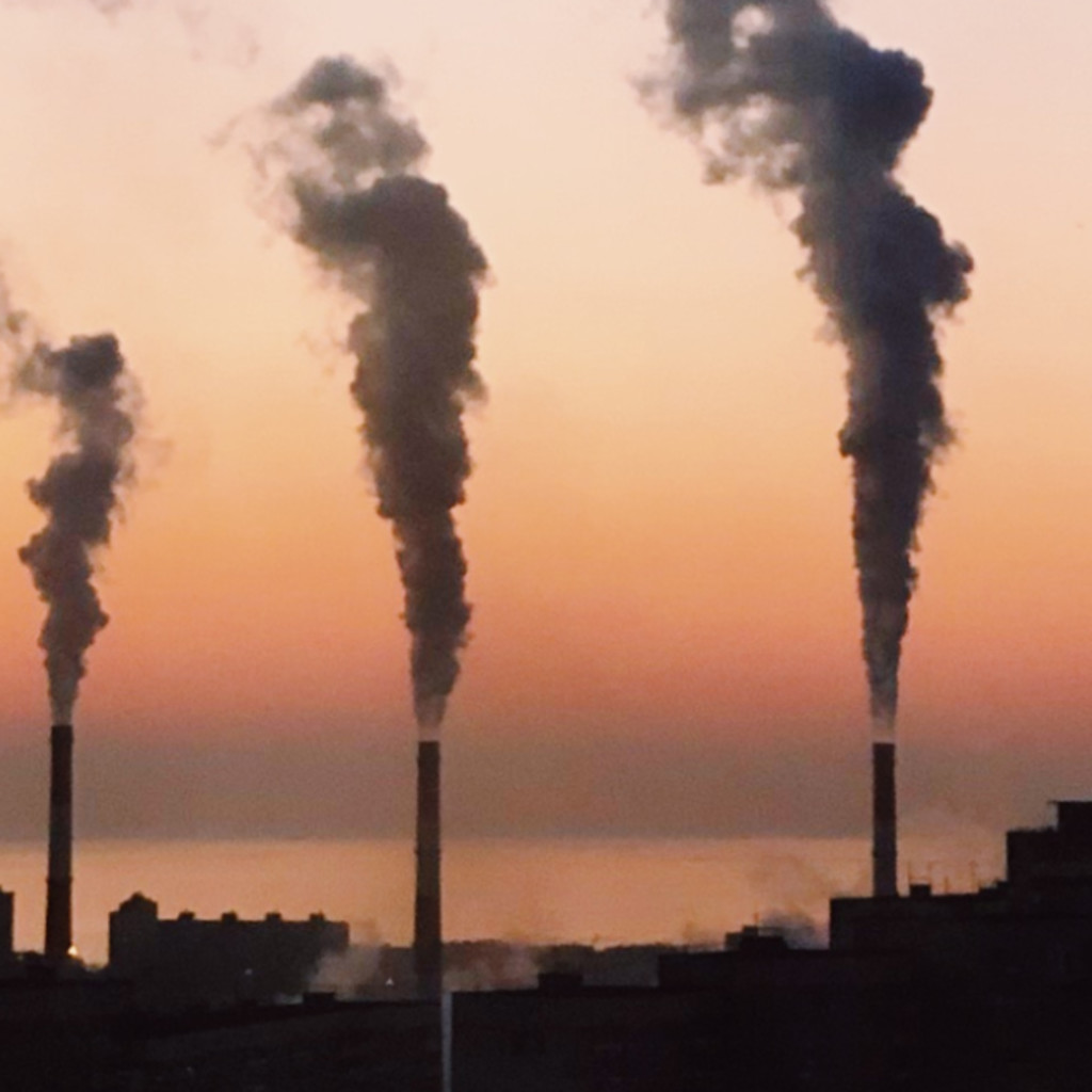 Factory smokestacks emit thick smoke against a colourful sunset sky, depicting pollution and industrial impact on the environment.