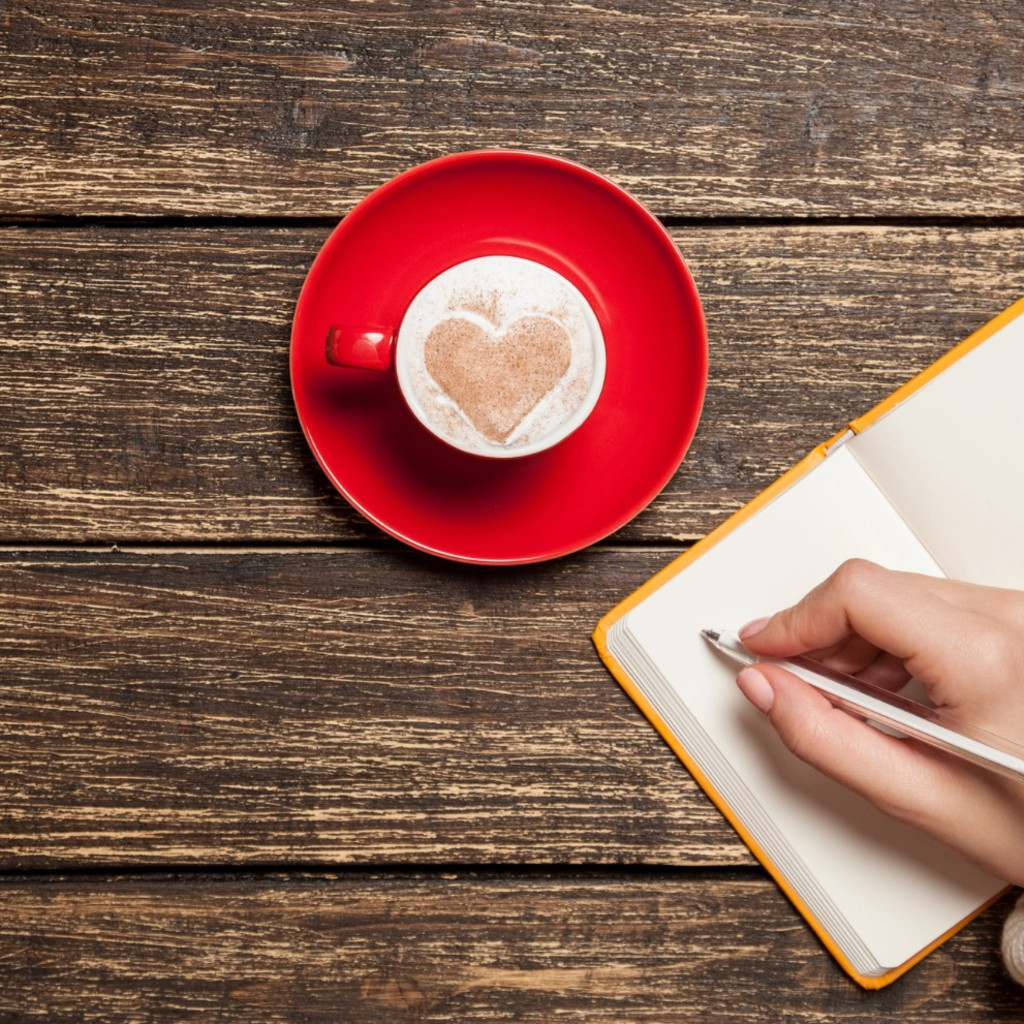 wooden background with a red cup and a heart in the foam, there is a notebook with a hand writing on the left