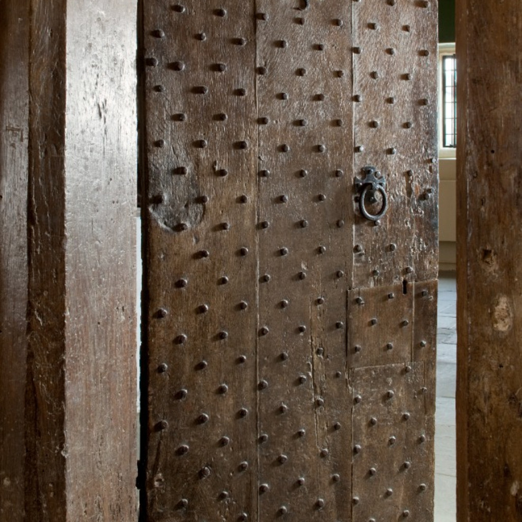 Picture of an old wooden door, with thick wooden surround. The door is studded with wooden pegs, and has a black iron circular handle. The door is slightly ajar.