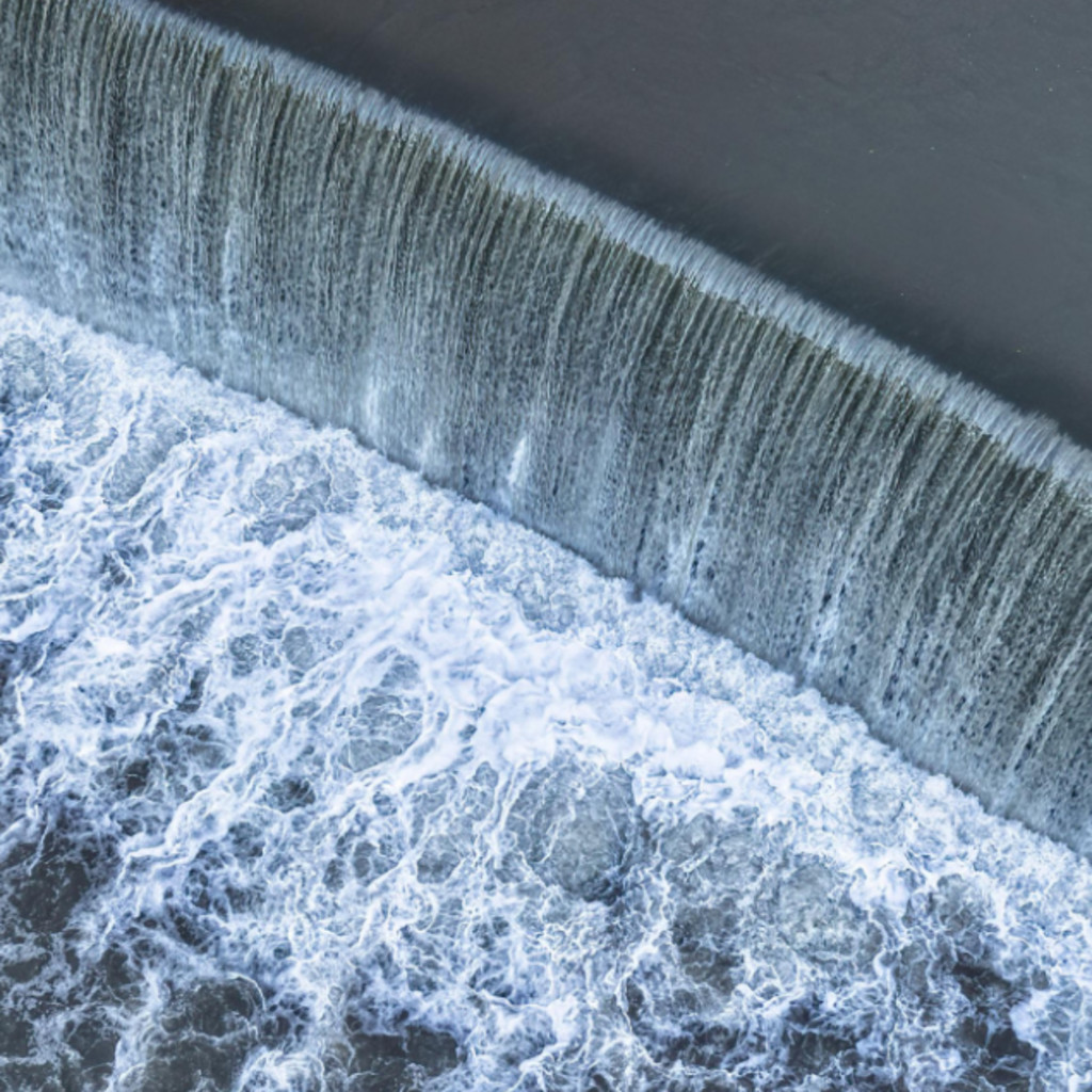 Aerial view of a curved dam with water cascading over, creating white foam.