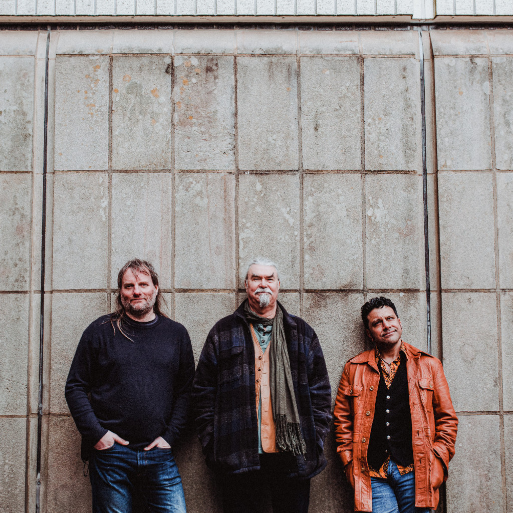 The image features Roger Flack, Peter Knight and Sacha Trochet wearing casual clothes, standing, leaning against a concrete wall