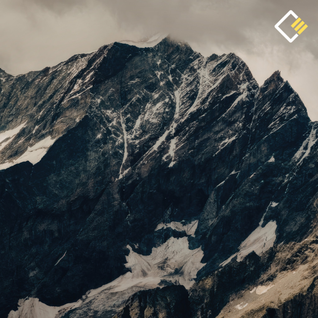Snow-dusted rugged mountain peak under a cloudy sky, with dramatic dark rock formations. The scene conveys a sense of solitude and awe.