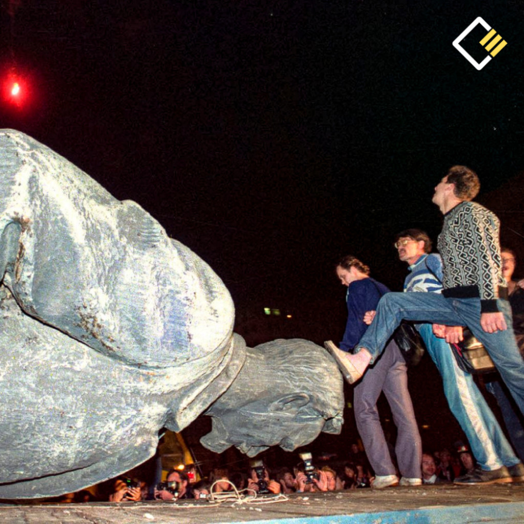 A group of people walks across a toppled statue of Lenin at night, one man stepping on its head. The scene conveys defiance and change.