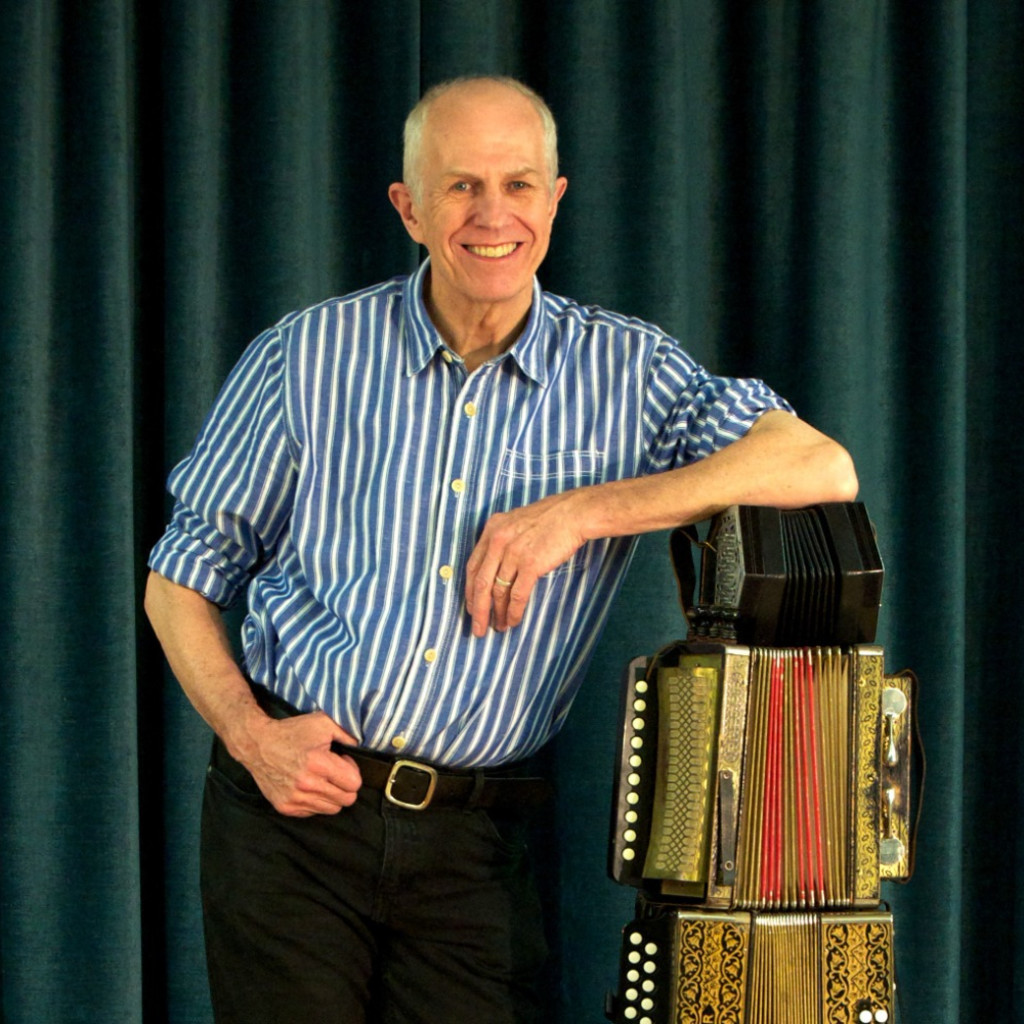 The image features a man wearing a blue and white pinstriped shirt and black trousers, standing next to two melodeons and a concertina. His left arm is resting on top of the instruments.