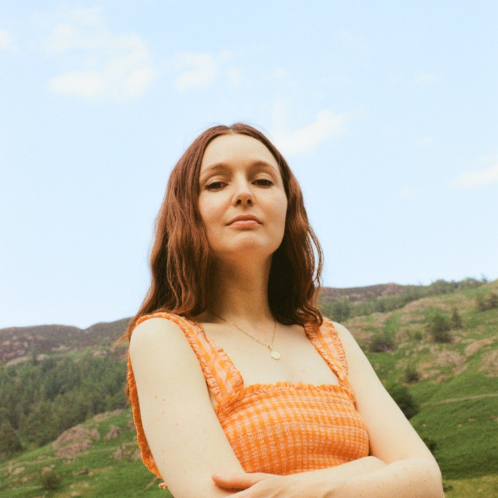 A young woman stands in a landscape hills. She has ginger hair and an orange top, and is posing with her arms folded.