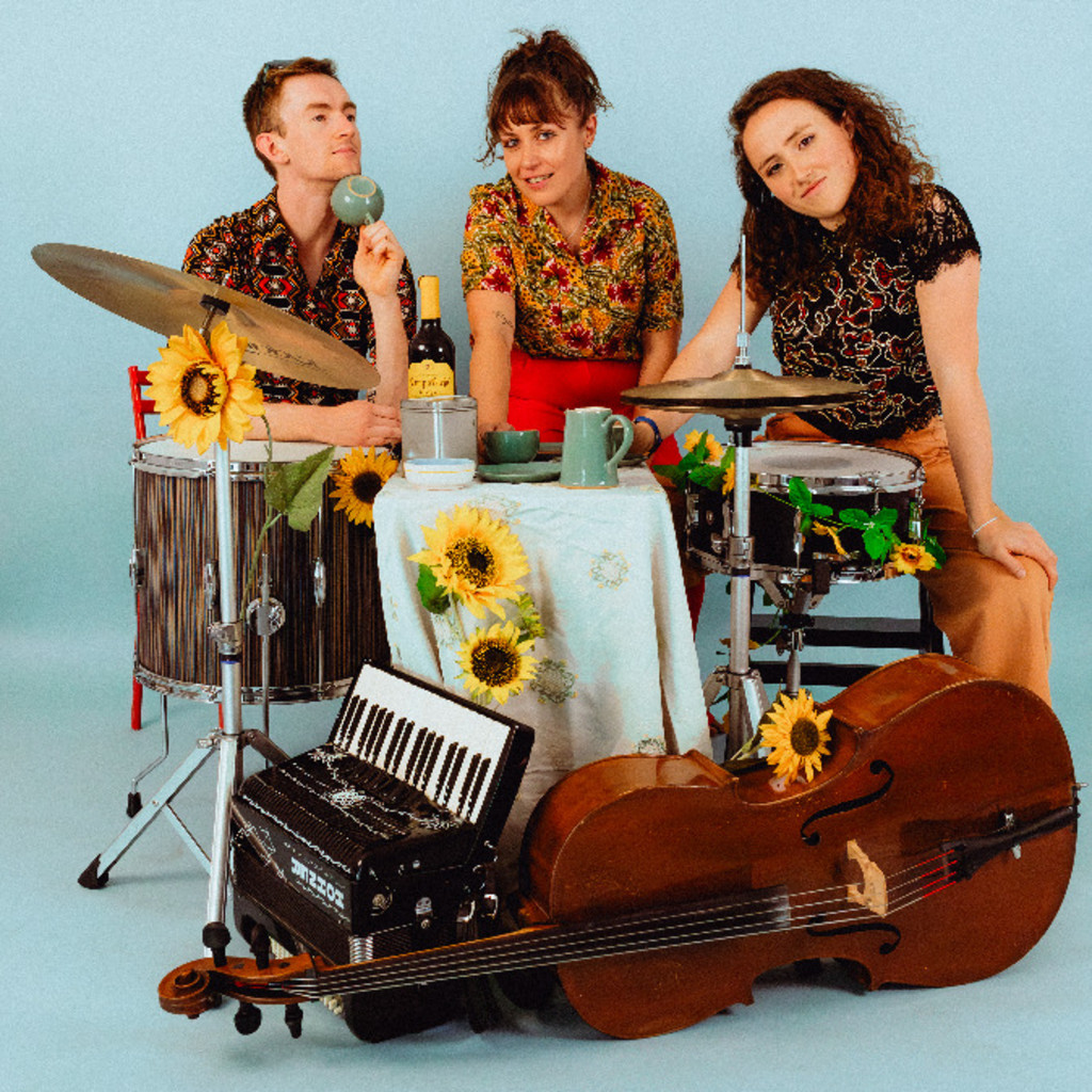 The image features Petee Shaw, Jenny Walinetski and Bonnie Swartz sitting behind a drum kit with an accordian and chello lying in front. The instruments have sunflowers lying across them.
