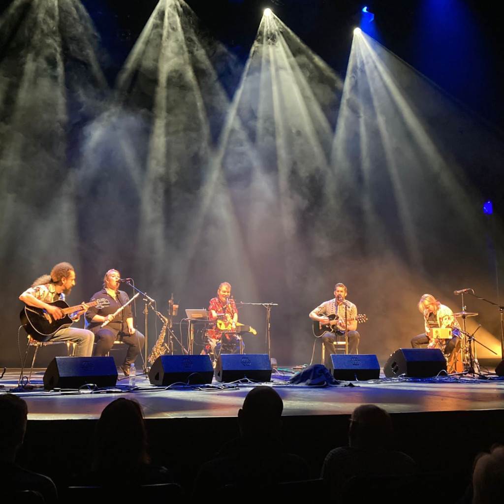 The image features John Kelly and his band sitting on stage performing with their instruments under overhead spotlights