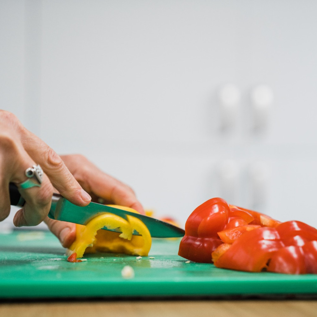 A woman's hand slicing yellow and red peppers on a green chopping board.
