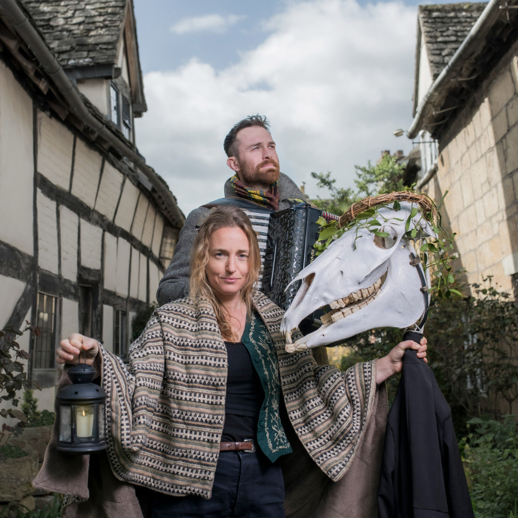 The image features Hannah Moore and Danny Pedler stood in a narrow street with old houses on either side. Hannah is holding a lantern in one hand and a horse's skull in the other