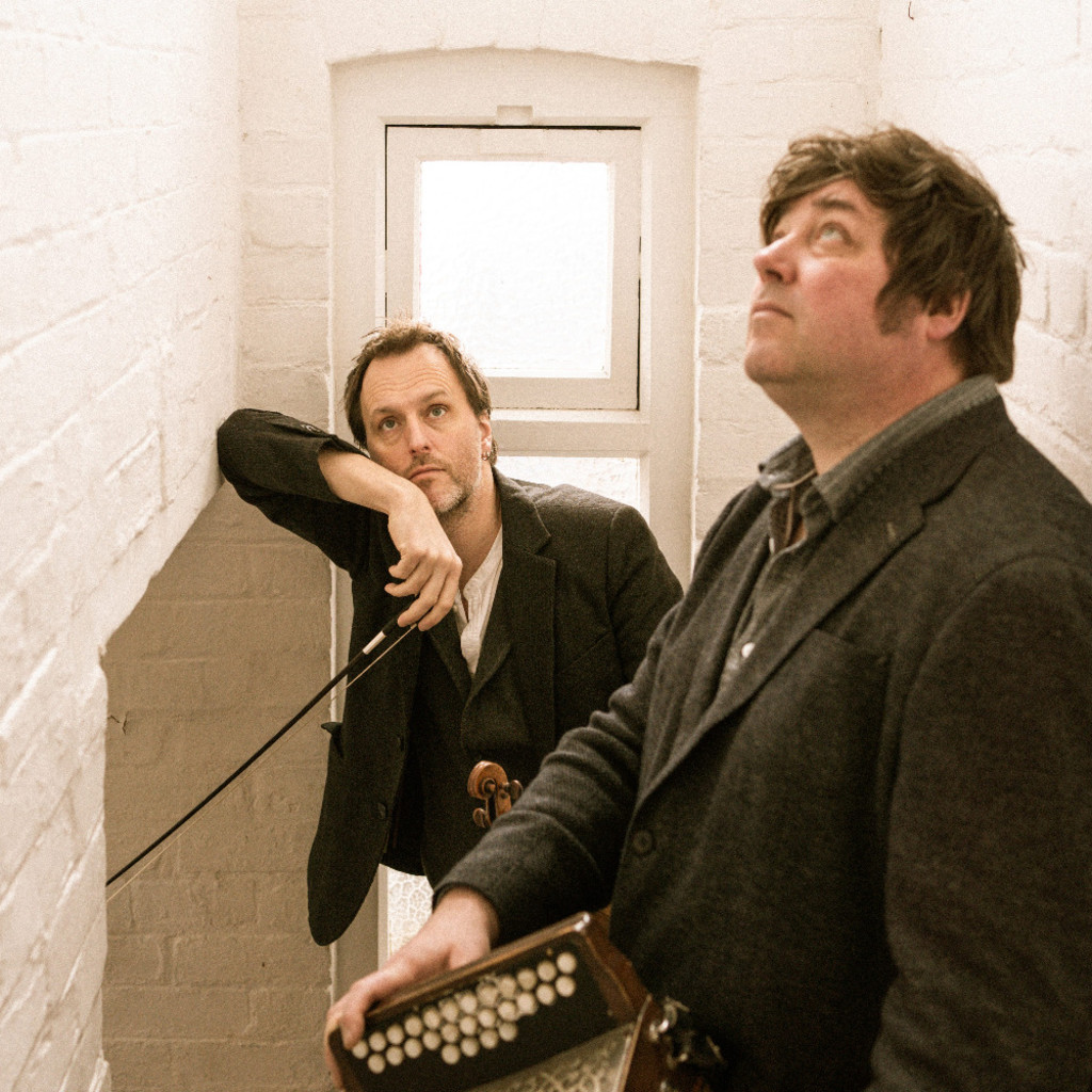 The image features John Spiers and Joe Boden in a white walled hall, in front of a small window, leaning against opposite walls and holding their instruments. They are looking up at the ceiling.