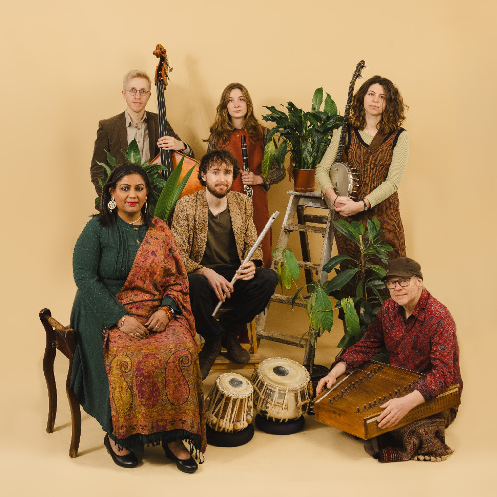 The image features the band Mishra and Deepa Shakthi sitting and standing in an ensemble holding thier instruments, with some potted plants in front of a pale orange background