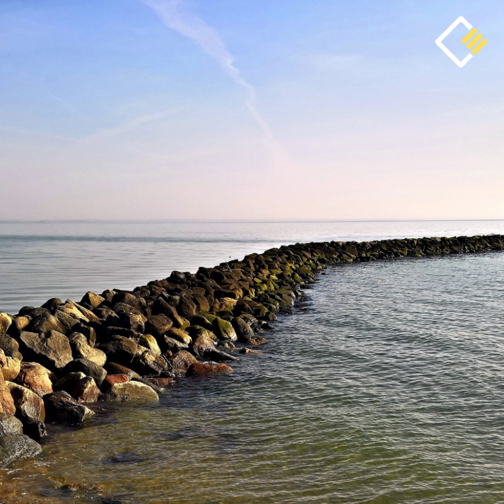 Curved rock barrier extends into calm sea under a blue sky, creating a serene, peaceful coastal scene. Cooper Education logo appears on the top right hand side.